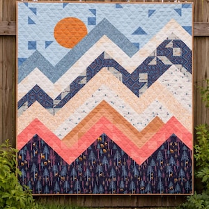 May include: A handmade quilt with a mountain landscape design. The quilt features a blue sky with an orange sun, layered mountain peaks in shades of blue, peach, and coral, and a forest scene at the base.