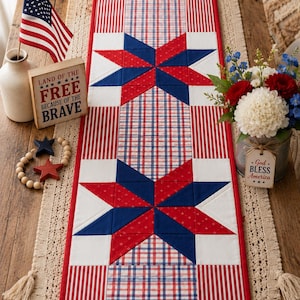 May include: A rectangular quilted table runner featuring a patriotic design with two large eight-pointed stars in red and blue, surrounded by red and white striped fabric and a red, white, and blue plaid pattern, all bordered by a solid red fabric trim.
