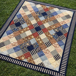 Op de afbeelding: Een patchwork quilt met vierkanten in verschillende patronen in blauw, rood, beige en bruin. De quilt heeft een marineblauwe rand met witte strepen en ligt op groen gras.