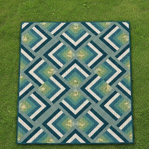 May include: A square quilt with a geometric pattern of teal, blue, and white chevron shapes. The quilt has a repeating pattern with a floral design in the center of each diamond. The quilt is on a green grassy surface.