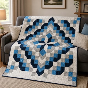 May include: A geometric patterned quilt in shades of blue, gray, and white, draped over a gray sofa. The quilt features a repeating block design with a central starburst pattern. The quilt's dimensions are not visible in the image.