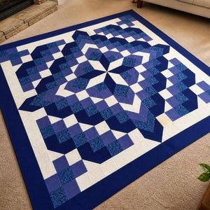 May include: A square quilt with a geometric design in shades of blue and white. The quilt features a central starburst pattern surrounded by blocks of varying shades of blue and white, with a solid blue border. The quilt is approximately 60 inches by 60 inches.