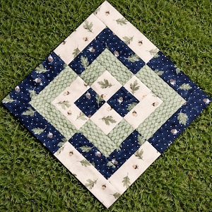 May include: A square quilted placemat with a diamond pattern. The design features alternating bands of navy blue with white polka dots and acorn and leaf prints, cream with acorn and leaf prints, and light green with a crosshatch pattern. The placemat is on a green grass background.