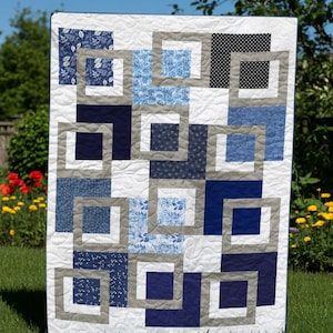 May include: A handmade quilt with a geometric design. The quilt features squares in various shades of blue, white, and gray, arranged in a repeating pattern. The squares are framed by gray borders, creating a modern and stylish look. The quilt is displayed outdoors on a sunny day.
