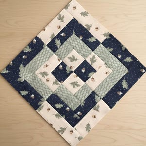 Könnte beinhalten: Ein quadratischer Patchwork-Quilt mit einem sich wiederholenden Muster aus Eichenblättern und Eicheln. Das Design umfasst marineblaue, cremefarbene und hellgrüne Stoffe. Der Quilt ist in einer Raute angeordnet und zeigt die komplizierten Nähte und Stoffauswahlen.