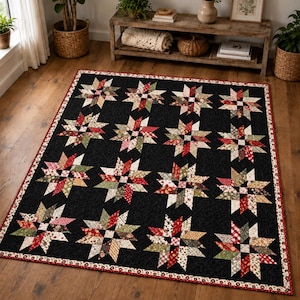 Op de afbeelding: Een rechthoekige quilt met een herhalend patroon van veelkleurige sterblokken in tinten rood, groen en crème, geplaatst tegen een effen zwarte achtergrond. De quilt is afgewerkt met een dunne rood-witte bloemenrand.