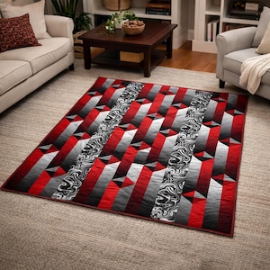 May include: A geometric patterned quilt in red, black, white, and gray. The design features repeating shapes and a marbled stripe. The quilt is rectangular and lies on a neutral-colored rug.