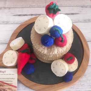 May include: A felt food play set with a stack of pancakes topped with white felt circles, red strawberries, and blue and red felt berries. Additional felt food items include red peppers and white felt slices. The set is displayed on a wooden serving board.