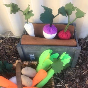 May include: A wooden crate filled with felt vegetables, including a white turnip, a purple and white turnip, and a red beetroot. The crate is sitting on a brown surface with green leaves and a basket of carrots and green vegetables.