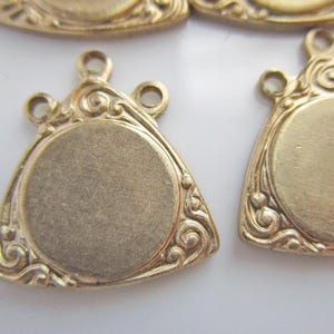 May include: Close-up of several gold-colored metal pendants. Each pendant has a triangular shape with a circular center and ornate scrollwork around the edges. The pendants have small loops at the top for attaching to a chain or other jewelry component.