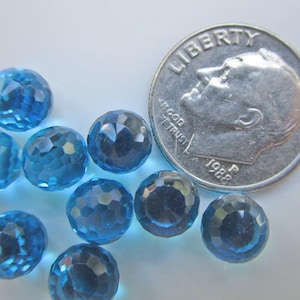 May include: A collection of faceted, round, light blue gemstones. A U.S. dime from 1988 is next to the gemstones for size comparison. The gemstones have a sparkling, crystal-like appearance.