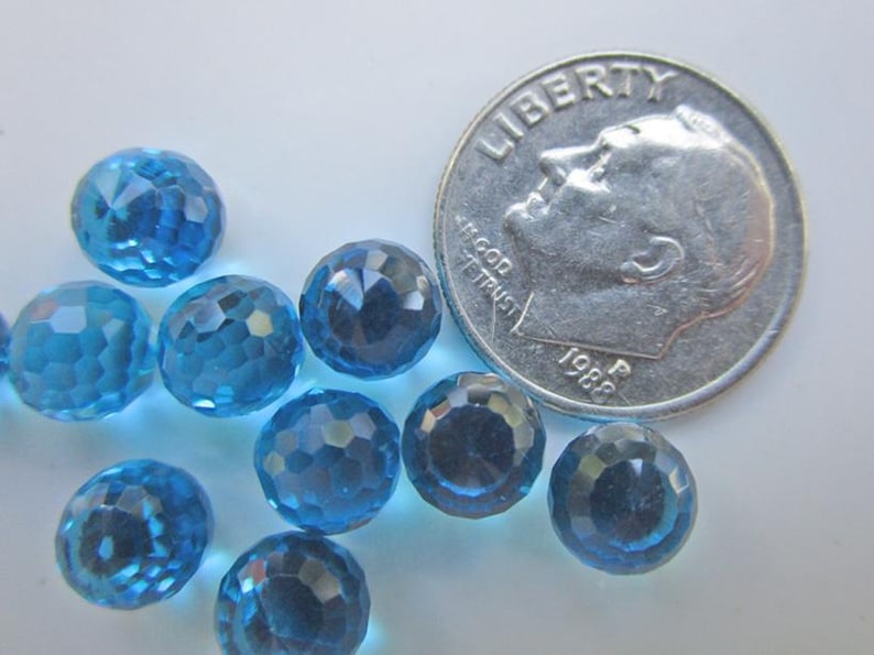 May include: A collection of faceted, round, light blue gemstones. A U.S. dime from 1988 is next to the gemstones for size comparison. The gemstones have a sparkling, crystal-like appearance.