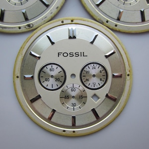 May include: Close-up of several Fossil watch faces. The watch faces are cream-colored with black numerals and hands. The word "FOSSIL" is printed in black. The faces have three smaller dials and a gold-colored outer ring.