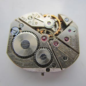 May include: A detailed view of a vintage Croton watch movement. The silver mechanism displays intricate gears, jewels, and the text "SEVENTEEN JEWELS". The watch movement is marked "UNADJUSTED" and "SWISS".