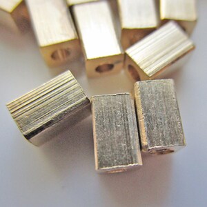 May include: Close-up of several small, rectangular brass beads. Each bead has a hole running through the center and a textured surface. The beads are scattered on a light background.