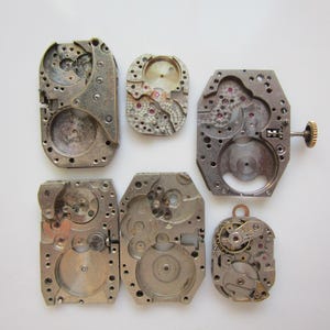 May include: A selection of antique watch movements, displaying intricate mechanical parts. The silver metal components feature gears, screws, and other internal elements, with some including red jewels. The arrangement highlights the detailed craftsmanship of vintage timepieces.