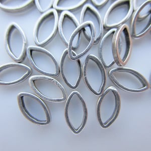 May include: A collection of small, silver-colored, oval-shaped metal beads. The beads have a hollow, open design and are scattered across a light-colored surface. These components are suitable for jewelry making.
