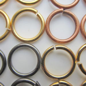 May include: Close-up of various metal jump rings in gold, copper, and gunmetal gray. The rings are open, ready for use in jewelry making or other crafts. The rings are arranged in rows against a white background.