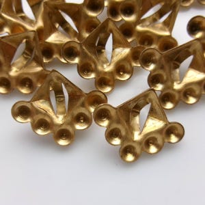 May include: Close-up of several small, ornate, gold-colored metal findings. Each finding has a triangular shape with a central opening and rounded, petal-like details. The findings are scattered on a white surface.