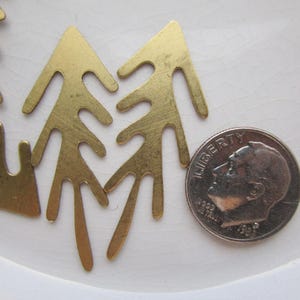 May include: Several small, brass-colored metal tree-shaped charms are arranged on a white surface next to a U.S. dime. The charms have a cutout design and are likely for jewelry making or crafting.