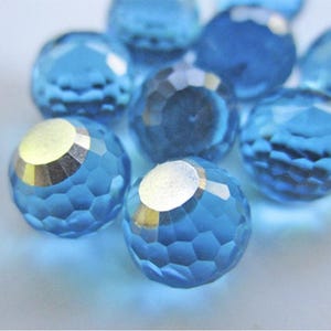May include: Close-up of several faceted, round, blue crystal beads. Each bead has a silver-colored metallic cap. The beads are scattered on a light blue surface, creating a sparkling effect. Ideal for jewelry making.