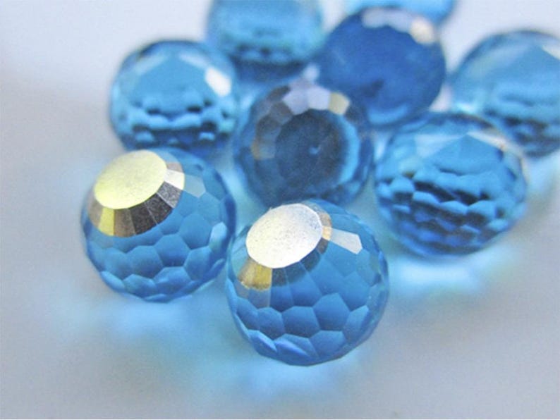 May include: Close-up of several faceted, round, blue crystal beads. Each bead has a silver-colored metallic cap. The beads are scattered on a light blue surface, creating a sparkling effect. Ideal for jewelry making.