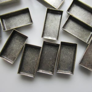 May include: A collection of rectangular, antique silver-toned metal bezels. The bezels are empty, with a shallow, open design, and are scattered across a white surface. These components are likely for jewelry making or crafting.