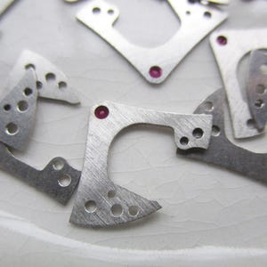 May include: Close-up of several small, silver metal parts with holes and rounded edges. Some of the parts have small, pink gemstones attached.