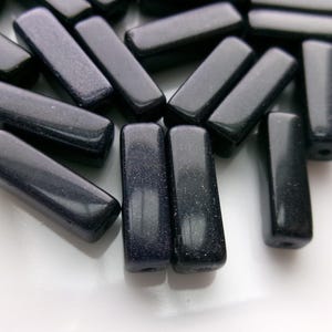 May include: A collection of dark blue rectangular beads with a sparkling, speckled appearance. The beads are smooth and polished, with rounded edges and a hole drilled through the center for stringing. These beads are suitable for jewelry making.