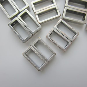 Könnte beinhalten: Nahaufnahme mehrerer rechteckiger Metallschnallen mit einem antiken, silberfarbenen Finish. Die Schnallen sind auf einer weißen Oberfläche angeordnet und zeigen ihr offenes, rechteckiges Design. Ideal für Bastelarbeiten und Schmuckherstellung.