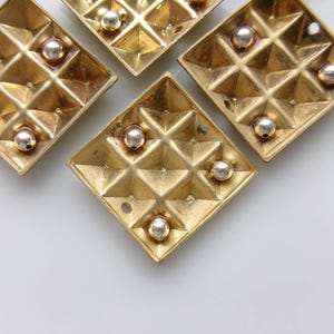 May include: Close-up of five gold-colored, diamond-shaped decorative elements. Each element features a grid pattern and small silver-colored spheres. The elements are arranged against a white background.
