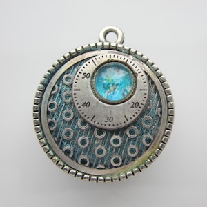 May include: A round, silver-toned pendant with a textured teal background and a central dial design. The dial features numbers and a small, round, blue and green center. The pendant has a loop at the top for hanging.