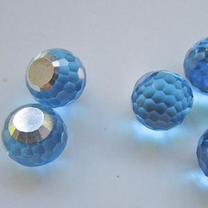 May include: Several faceted, light blue crystal beads with a silver-colored backing. The beads have a spherical shape with a geometric pattern of small facets. The beads are scattered on a white surface.