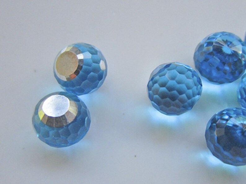 May include: Several faceted, light blue crystal beads with a silver-colored backing. The beads have a spherical shape with a geometric pattern of small facets. The beads are scattered on a white surface.