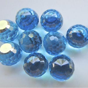 May include: A collection of faceted, light blue crystal beads. Each bead has a spherical shape with multiple cut surfaces, creating a sparkling effect. The beads are scattered on a white surface, reflecting light.