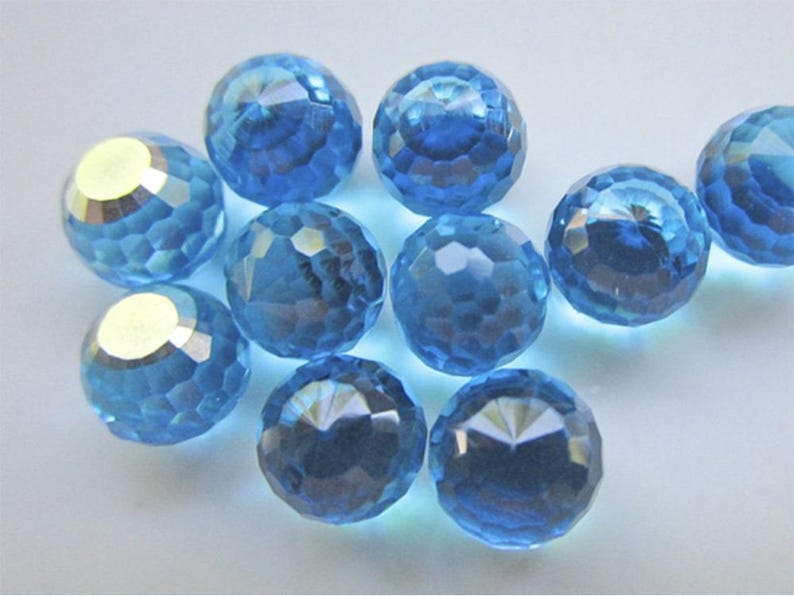 May include: A collection of faceted, light blue crystal beads. Each bead has a spherical shape with multiple cut surfaces, creating a sparkling effect. The beads are scattered on a white surface, reflecting light.