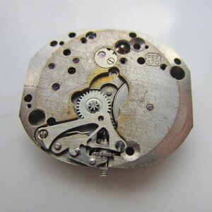 May include: Detailed view of a watch mechanism, showcasing small gears, screws, and metal components. The watch parts are silver and gold in colour. The text "FEF 170" is stamped on the metal.