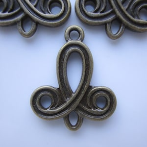 May include: Close-up of antique bronze-colored metal embellishments. Each piece features a central loop design with decorative swirls at the bottom. The embellishments have a small loop at the top for attachment. Ideal for jewelry making or craft projects.