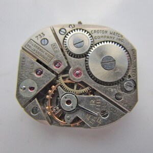 May include: A detailed view of a vintage Croton watch movement. The silver mechanism displays various gears, jewels, and the text "CROTON WATCH COMPANY INC." and "SEVENTEEN JEWELS". The watch movement is non-magnetic and Swiss made.