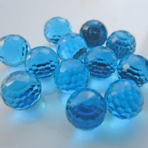 May include: A collection of faceted, round, light blue glass beads. The beads have a cut-glass appearance, reflecting light and creating a sparkling effect. The beads are scattered on a white surface.