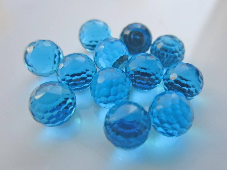 May include: A collection of faceted, round, light blue glass beads. The beads have a cut-glass appearance, reflecting light and creating a sparkling effect. The beads are scattered on a white surface.