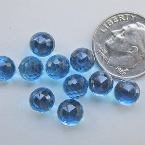 May include: A collection of faceted, round, blue gemstones. A U.S. dime is included for size comparison. The gemstones have a sparkling, multi-faceted surface. The color is a deep, transparent blue. The gemstones are scattered on a light-colored surface.