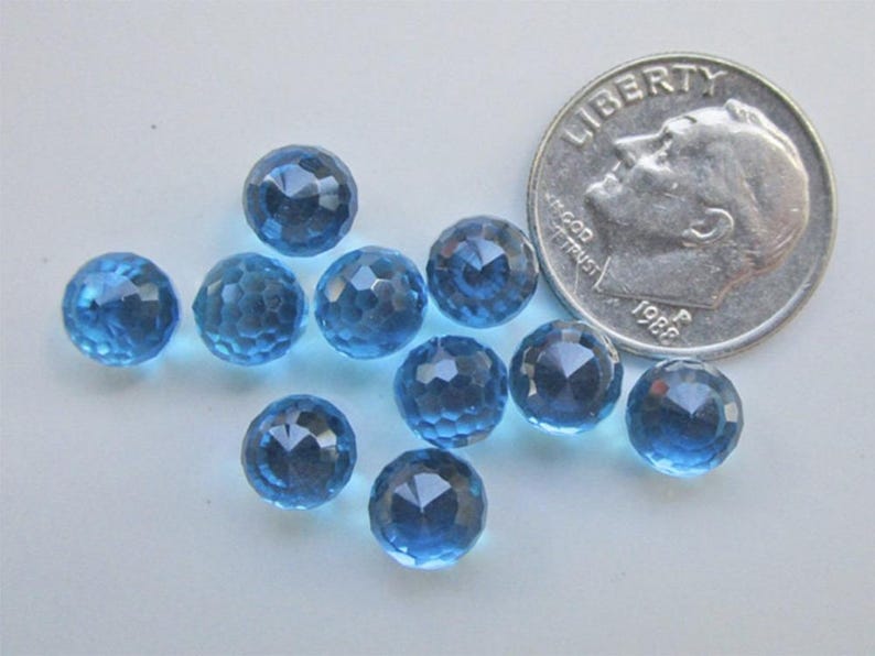 May include: A collection of faceted, round, blue gemstones. A U.S. dime is included for size comparison. The gemstones have a sparkling, multi-faceted surface. The color is a deep, transparent blue. The gemstones are scattered on a light-colored surface.
