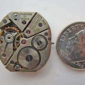 May include: Detailed image of a vintage watch movement, showcasing numerous gears, jewels, and text. A UK ten pence coin is placed beside it for scale. The watch mechanism has text that reads "SEVENTEEN 17 JEWELS" and "CROTON WATCH COMPANY INC."