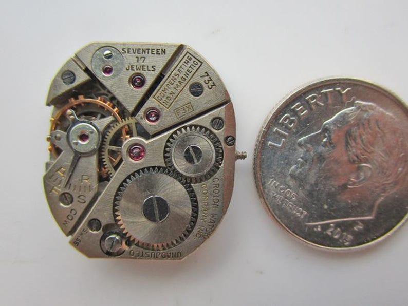 May include: Detailed image of a vintage watch movement, showcasing numerous gears, jewels, and text. A UK ten pence coin is placed beside it for scale. The watch mechanism has text that reads "SEVENTEEN 17 JEWELS" and "CROTON WATCH COMPANY INC."