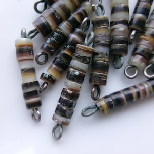 May include: Close-up of several strands of beads. The beads are cylindrical, with alternating bands of dark brown, tan, and white. Each bead has a metal loop attached for stringing. The beads are scattered on a white surface.