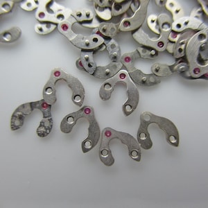 May include: A close-up of a pile of small, silver-coloured metal pieces with a U-shaped design. Each piece has a small, pink jewel in the centre.