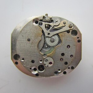 May include: A detailed view of a vintage watch movement, showcasing a complex arrangement of gears, levers, and jewels. The metal components are silver-toned, with numerous small holes. The watch mechanism is a complex assembly of tiny parts.