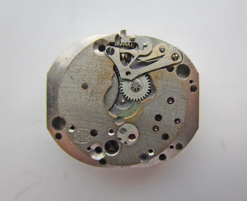 May include: A detailed view of a vintage watch movement, showcasing a complex arrangement of gears, levers, and jewels. The metal components are silver-toned, with numerous small holes. The watch mechanism is a complex assembly of tiny parts.