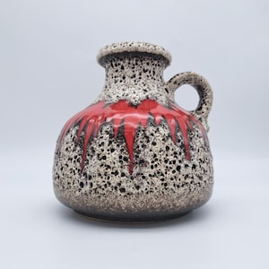 May include: A ceramic vase with a handle, featuring a textured, speckled surface in shades of brown, black, and cream. The vase has a vibrant red glaze that drips down the sides, adding a pop of colour to the design.